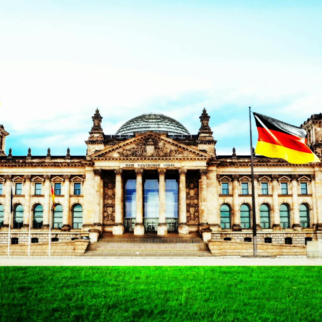 Bild des Bundestag-/Reichtagsgebäudes in Berlin mit Detuschlandfahne und Rasen davor.